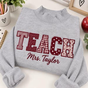 May include: A light gray crewneck sweatshirt featuring the word TEACH in large, maroon-colored block letters with varied patterns including stripes, polka dots, gingham, stars, and floral prints, with the name Mrs. Taylor embroidered below.