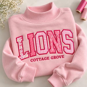 May include: A light pink mock neck sweatshirt featuring the word LIONS in large, patterned applique letters with pink floral, polka dot, gingham, and striped designs. Below the word LIONS, the text COTTAGE GROVE is embroidered in dark pink.