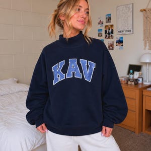 May include: A navy blue mock neck sweatshirt featuring the letters KAV in a white-bordered, blue and white gingham check pattern across the chest. The garment has long sleeves with ribbed cuffs and a ribbed hem, providing a relaxed fit.
