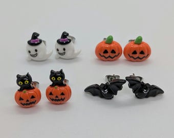 May include: A collection of Halloween-themed stud earrings. The earrings feature designs of ghosts in witch hats, orange pumpkins, black cats on pumpkins, and black bats. The earrings are set against a white background.