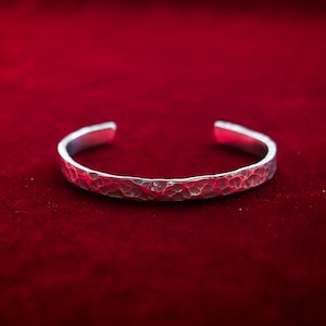May include: A silver cuff bracelet with a hammered texture, set against a deep red background. The open design allows for easy wear. This bracelet is a simple yet elegant accessory.