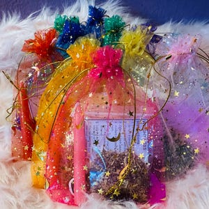 May include: A selection of colourful organza bags, each filled with different items. The bags are tied with matching ribbons and decorated with gold stars. Some bags contain small candles and dried herbs. The background is a fluffy, light pink material.