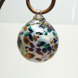 May include: A small, round, white glass ornament with a variety of colorful flecks and swirls. The ornament is hanging from a copper hook.