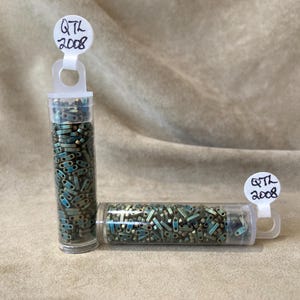 May include: Three clear plastic tubes filled with small, teal and bronze-colored beads. Each tube has a white tag with "QTL 2008" written on it. The tubes are arranged in an L-shape against a neutral background.