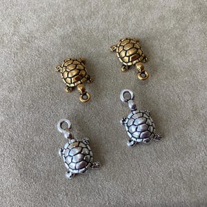 May include: Four small turtle charms in gold and silver tones. The charms have detailed shell patterns and small loops for attaching to jewelry. The turtles are arranged on a neutral-colored surface.