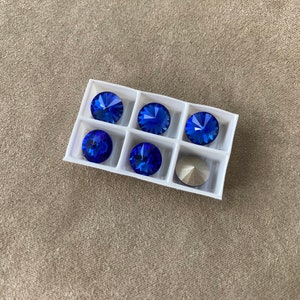 May include: A white plastic tray with six blue rhinestones and one silver rhinestone. The rhinestones are arranged in a grid pattern.