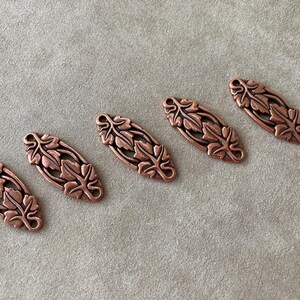 May include: Five copper-colored metal leaf-shaped embellishments. Each has a detailed leaf design and two small holes at each end. The embellishments are arranged diagonally on a neutral-toned surface.