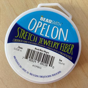 May include: A spool of black stretch jewelry fiber, labeled "Beadsmith Opelon Stretch Jewelry Fiber" with the text "Cordon de Fibre Elastique a Bijoux - Hbra Elastica para Bisuteria". The spool is 5 meters (16.4 feet) long and 0.7 mm (0.028 inches) thick.