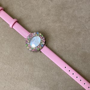 May include: A pink wristwatch with a light pink leather band. The watch face is surrounded by a halo of colorful gemstones. The watch has a silver buckle.