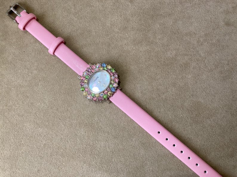May include: A pink wristwatch with a light pink leather band. The watch face is surrounded by a halo of colorful gemstones. The watch has a silver buckle.