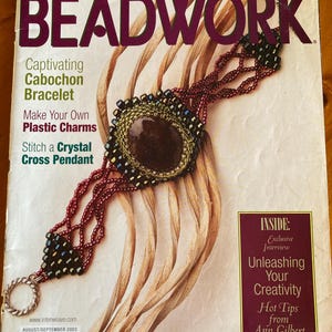 May include: A magazine cover for "BEADWORK" displays a beaded bracelet with a large, oval, brown cabochon. The bracelet is red and black, with a silver clasp. The magazine cover has text that reads "Captivating Cabochon Bracelet".