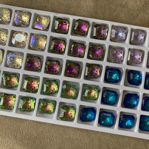 Op de afbeelding: Een witte lade met 36 heldere glazen strasssteentjes in verschillende kleuren, waaronder blauw, groen, paars en helder. De strasssteentjes zijn gerangschikt in rijen en kolommen, en elk strasssteentje heeft een uniek ontwerp.