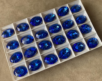 May include: A close-up view of a white tray filled with 24 oval-shaped, blue glass rhinestones. The rhinestones are arranged in rows and columns.