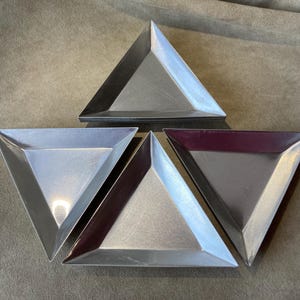 May include: Four silver-colored, triangular plates. The plates are arranged in a star-like pattern, with three plates facing outward and one in the center. One plate has a burgundy interior. The plates are made of metal and have a shiny finish.