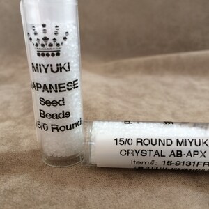 May include: Two small clear plastic tubes filled with white seed beads. The label on the larger tube reads "MIYUKI JAPANESE Seed Beads 15/0 Round". The label on the smaller tube reads "15/0 ROUND MIYUKI MATTE CRYSTAL AB-APX 8.20 M Item#: 15-9131FR-TE".