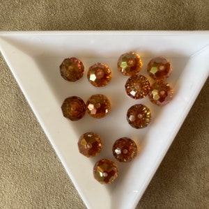 May include: A collection of round, faceted, iridescent glass beads in shades of amber and gold. The beads are arranged in a white triangular tray.