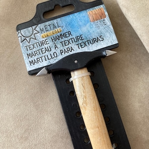 May include: A black and white texture hammer with a wooden handle. The packaging says "Metal Elements Texture Hammer" and "Marteau à Texture" and "Martillo Para Texturas".