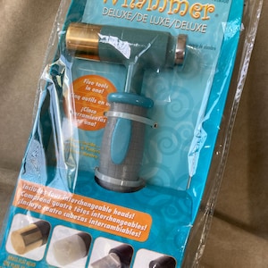 May include: The Whammer Deluxe multi-tool hammer in teal and grey packaging. The tool features a brass head and a textured grey handle. The packaging displays the text Five tools in one and Includes four interchangeable heads with images of brass, nylon, pick, and dapping heads.