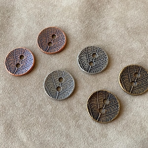 May include: Six round metal buttons with a leaf design. The buttons are in various colors, including copper, silver, and bronze. Each button has two holes for sewing. The buttons are on a tan surface.
