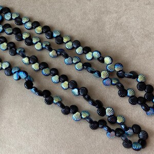 May include: Three strands of a necklace made of black shell-shaped beads with iridescent blue and yellow highlights. The beads are strung together with black string, creating a layered effect. The necklace is displayed on a neutral background.