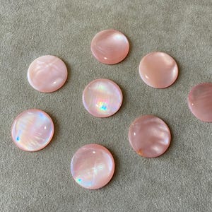 May include: Eight round, pink mother-of-pearl cabochons with iridescent highlights are scattered on a gray surface. The cabochons vary slightly in color, ranging from pale pink to a deeper rose hue. These decorative elements are ideal for jewelry making.