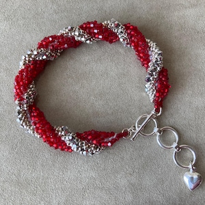 May include: A red and silver beaded bracelet with a heart-shaped charm. The bracelet is twisted and has a silver clasp.