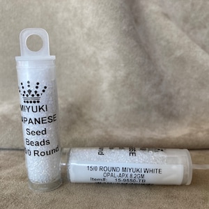 May include: Two small clear plastic containers filled with white seed beads. The container on the left has a black label that reads "MIYUKI JAPANESE Seed Beads 15/0 Round". The container on the right has a white label that reads "15/0 ROUND MIYUKI WHITE OPAL-APX 8.2GM Item#: 15-9550-TB".