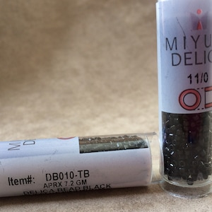 Peut inclure: Deux petits tubes en plastique transparent remplis de perles de rocaille noires. L'étiquette du tube de droite indique "MIYU DELIC 11/0". L'étiquette du tube de gauche indique "Item#: DB010-TB APRX 7.2 GM DELICA BEAD BLACK".