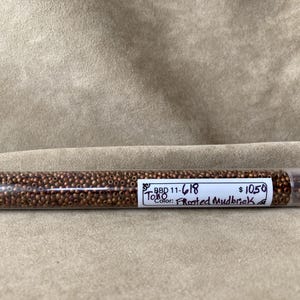 May include: A clear plastic tube filled with small, round, frosted brown beads. The tube has a white label with the text "BBD 11-618", "TORO", and "Color: Frosted Mudbrick". The beads are a warm, earthy tone.