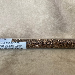May include: A clear plastic tube filled with small, bronze-colored, 11/0 cut Miyuki seed beads. The tube has a white label with the text "Item#:11C-9457-TB BRONZE(11C-614)APX21GM TB 11/0 CUT MIYUKI MET DK".