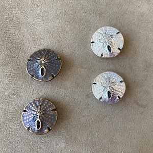 May include: Four decorative sand dollar shaped objects. Two are silver-colored with a detailed floral design, and two are bronze-colored with a similar design. The objects are round with a central hole and are likely used for home decor.