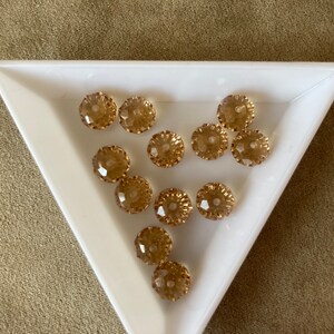 May include: A collection of 12 clear, faceted, round glass beads with a gold tone finish. The beads are arranged in a triangular tray.