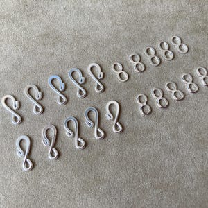 May include: A collection of silver-colored metal S-hooks and figure-eight shaped jewelry clasps arranged in two rows. These small components feature a smooth, reflective finish and are designed for connecting jewelry pieces or other small items.