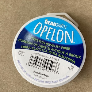 Puede incluir: Un carrete de fibra elástica negra Opelon para joyería. La etiqueta incluye el texto "STRETCH JEWELRY FIBER" y "FIBRA ELÁSTICA PARA BISUTERÍA". El carrete tiene 0,4 mm de grosor y contiene 5 m de fibra.
