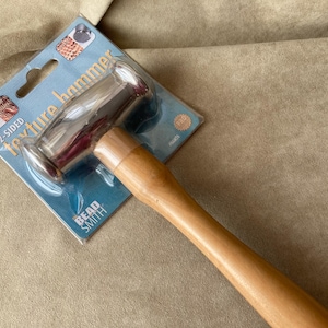 May include: A two-sided texture hammer with a wooden handle. The hammer has a silver head with two different textured surfaces. The packaging says "2-Sided Texture Hammer" and "Bead Smith".