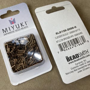 Może przedstawiać: Dwa opakowania japońskich koralików Miyuki. Jedno opakowanie przedstawia przezroczysty plastikowy pojemnik wypełniony smukłymi, brązowymi koralikami. Drugie opakowanie zawiera informacje o produkcie, w tym rozmiar koralika 1,3x6mm.