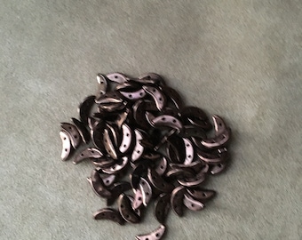May include: A pile of small, crescent-shaped metal beads in a dark bronze color. The beads have a smooth, shiny finish and are arranged in a random pattern.
