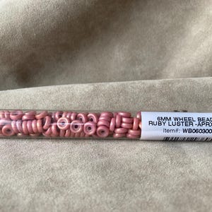 May include: A clear tube filled with approximately 19 grams of 6mm pink glass wheel beads. The label reads "6MM WHEEL BEAD CHALK RUBY LUSTER - APRX 19.GM/T Item#: WB060300-14497-T"