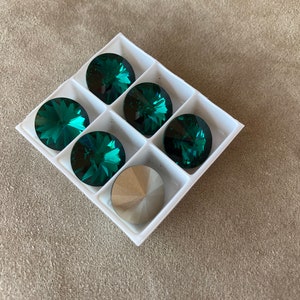 May include: A white plastic tray holding six faceted, emerald green crystal rhinestones and one silver rhinestone. The crystals are round with multiple facets, reflecting light. Ideal for jewelry making or embellishments.