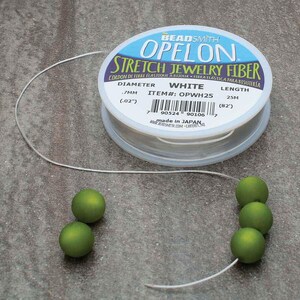 May include: A spool of white OPELON stretch jewelry fiber with a diameter of .7mm (.02"). The label also indicates a length of 25m (82'). Several green beads are strung on the fiber, showcasing its use in jewelry making.