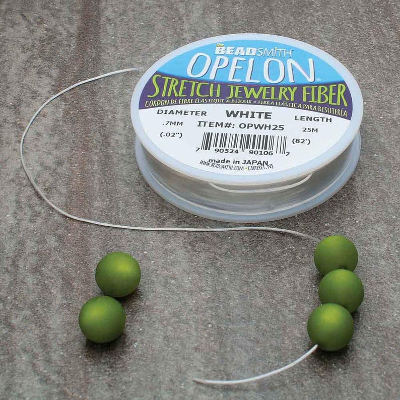 May include: A spool of white OPELON stretch jewelry fiber with a diameter of .7mm (.02"). The label also indicates a length of 25m (82'). Several green beads are strung on the fiber, showcasing its use in jewelry making.