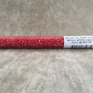 May include: A clear plastic tube filled with red 8/0 cut seed beads. The label reads "8/0 Cut S/L Ruby Hex Cut Miyuki Approx 20GM Tube Item#: 8C-911-TB".