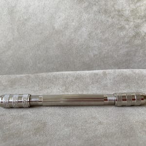 May include: A silver-toned metal pin vise tool featuring a central cylindrical handle with vertical fluting and two knurled, textured grip sections at each end. The tool has a polished metallic finish and a symmetrical, double-ended design.