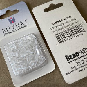 May include: Two packages of Miyuki Japanese seed beads. One package contains clear, slender bugle beads in a clear plastic container. The other package has product information, including the text "SLB136-420-R SLENDER BUGLE 1.3X6MM WHITE".