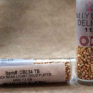 May include: Two small clear glass bottles filled with gold-colored seed beads. The bottle on the left has a label that reads "Item#: DB034-TB DELICA BEAD LT24KT GOLD PLATED APRX 7.2 3M". The bottle on the right has a label that reads "MIYUKI DELICA 11/0".