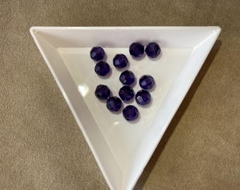 May include: A collection of faceted, dark purple beads are displayed in a white triangular dish. The beads have a glossy finish and are scattered across the dish. The background is a neutral tan color.