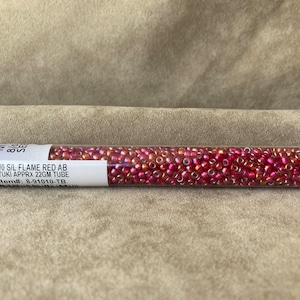 May include: A clear plastic tube filled with small, red, iridescent seed beads. The label on the tube reads "8/0 S/L FLAME RED AB MIYUKI APPRX 22GM TUBE Item#: 8-91010-TB".