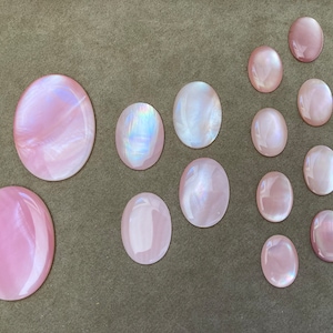 Può includere: Una collezione di 12 cabochon ovali iridescenti rosa e bianchi di diverse dimensioni. I cabochon sono disposti a semicerchio su una superficie marrone.
