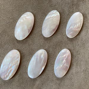 Könnte beinhalten: Sechs ovale Cabochons aus schillerndem Perlmutt. Die Cabochons haben einen perlmuttartigen Glanz mit zarten rosa- und weißen Farbtönen. Sie sind auf einer neutralen, strukturierten Oberfläche angeordnet, geeignet für Schmuckherstellung oder Bastelprojekte.
