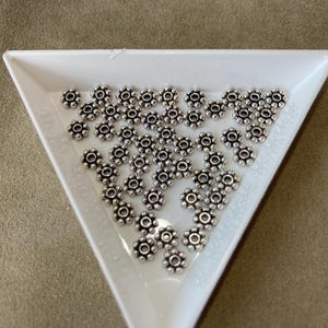 May include: A collection of small, silver-colored flower-shaped beads. The beads are arranged in a white, triangular tray against a neutral background. Each bead has a central hole and multiple petals.